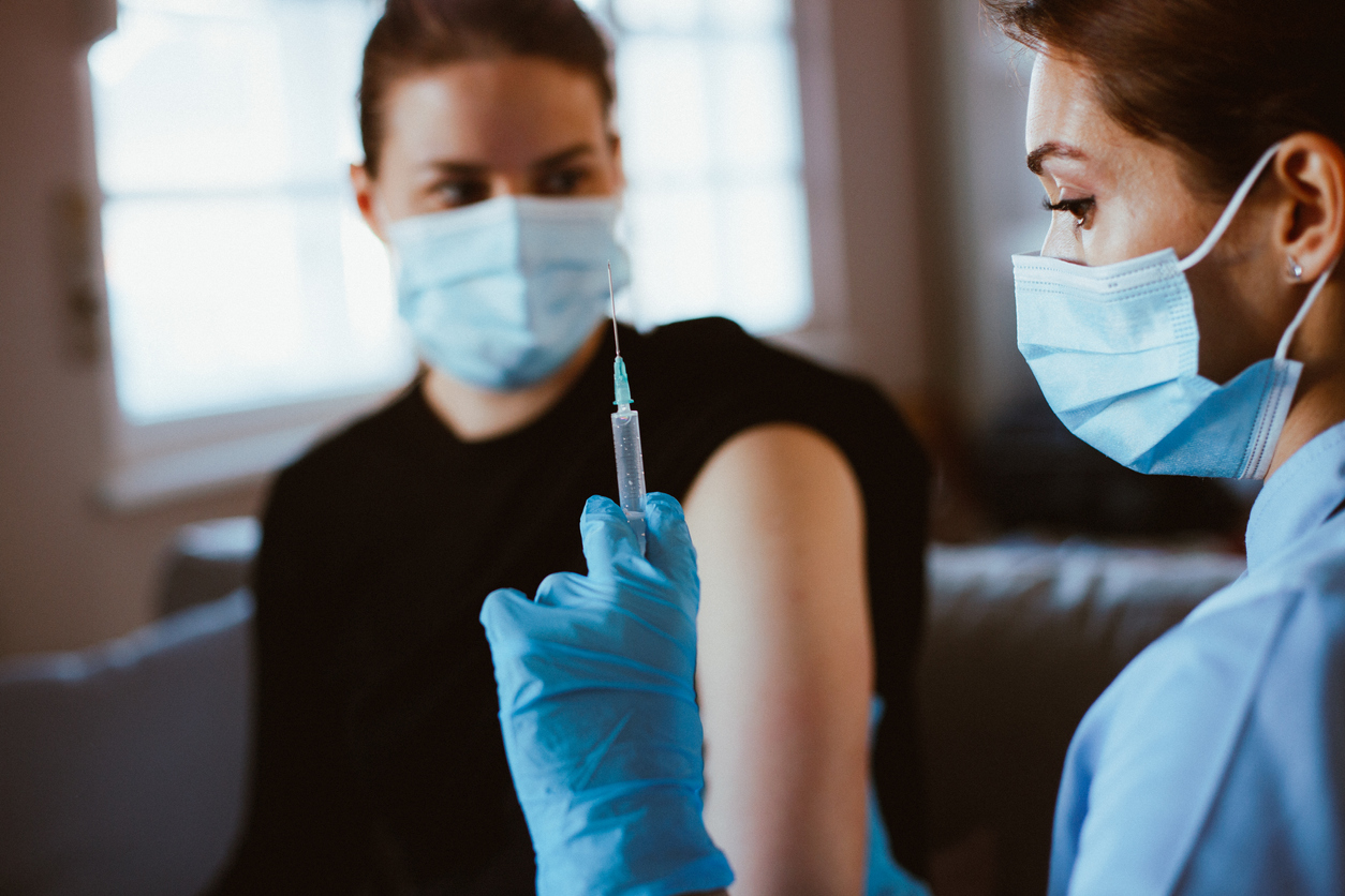 A young woman prepares to receive a COVID-19 vaccine from a female healthcare working holding a syringe and wearing a face mask and gloves.