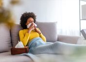 young woman in yellow sweater blowing nose on couch