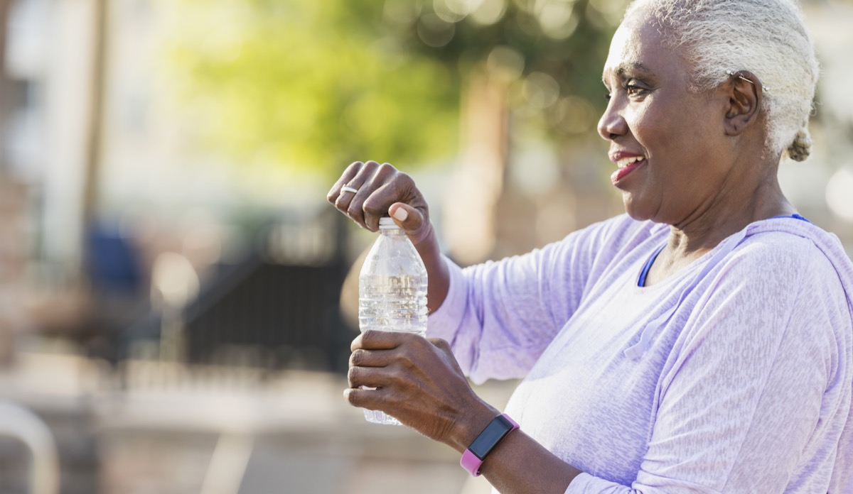 woman in her 60s wearing a fitness tracker and workout clothing, taking a break from exercising, opening a water bottle.