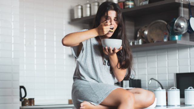 young woman eating bowl of cereal and fruit at home in kitchen.