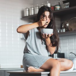 young woman eating bowl of cereal and fruit at home in kitchen.