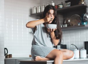 young woman eating bowl of cereal and fruit at home in kitchen.