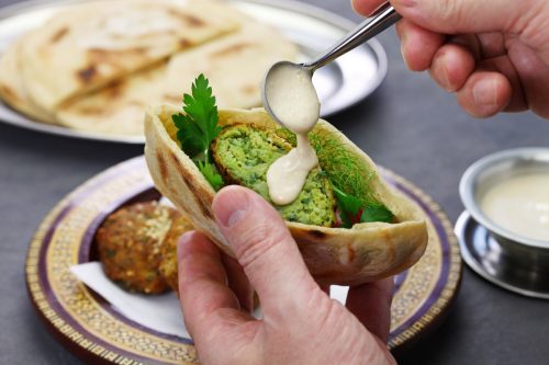 hand pouring tahini on falafel in a pita