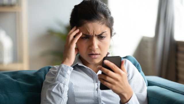 Confused young woman at home looking at cellphone having operational problems