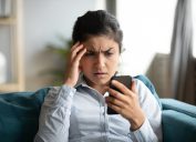 Confused young woman at home looking at cellphone having operational problems