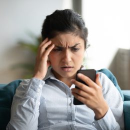 Confused young woman at home looking at cellphone having operational problems