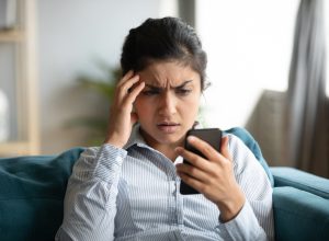 Confused young woman at home looking at cellphone having operational problems