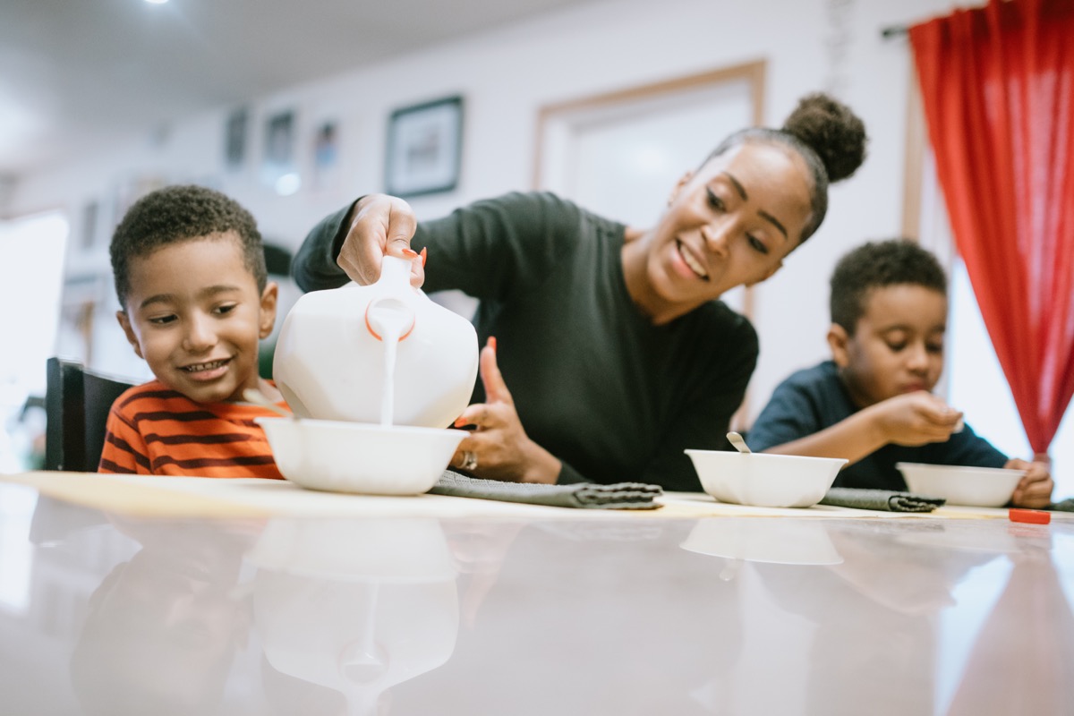 A mom sits at a table eating cereal with her sons at home. She pours milk into his bowl.
