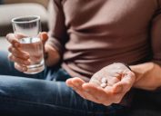 A person holding two pills in the palm of their hand and a glass of water