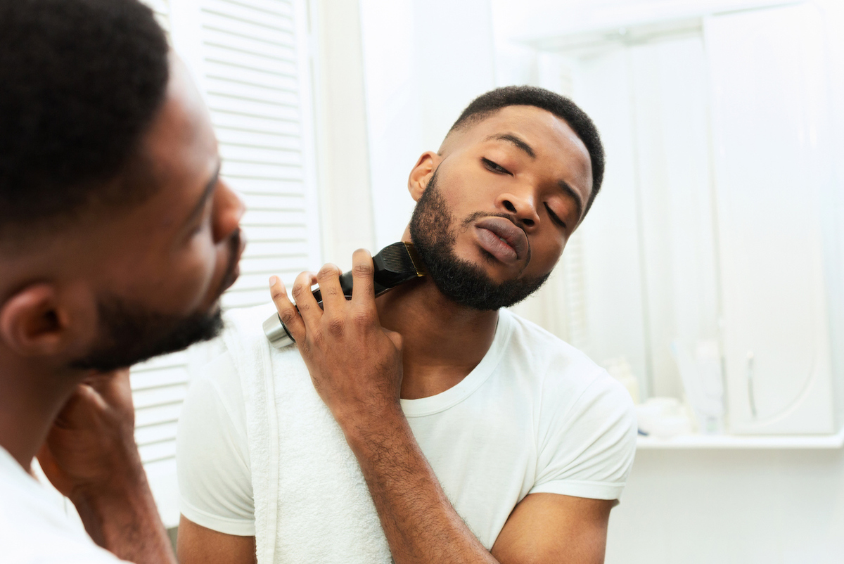 Young man looking at mirror and shaving beard with trimmer