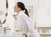 A young woman is standing in front of the bathroom mirror and brushing her teeth. Horizontally framed shot.