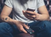 A young man is sitting on a sofa with a remote control and a smartphone