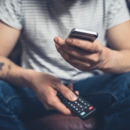 A young man is sitting on a sofa with a remote control and a smartphone