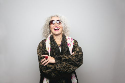 Cool mature woman wearing sunglasses, a fur coat, a floral scarf, and lot of jewelry, standing in front of grey background