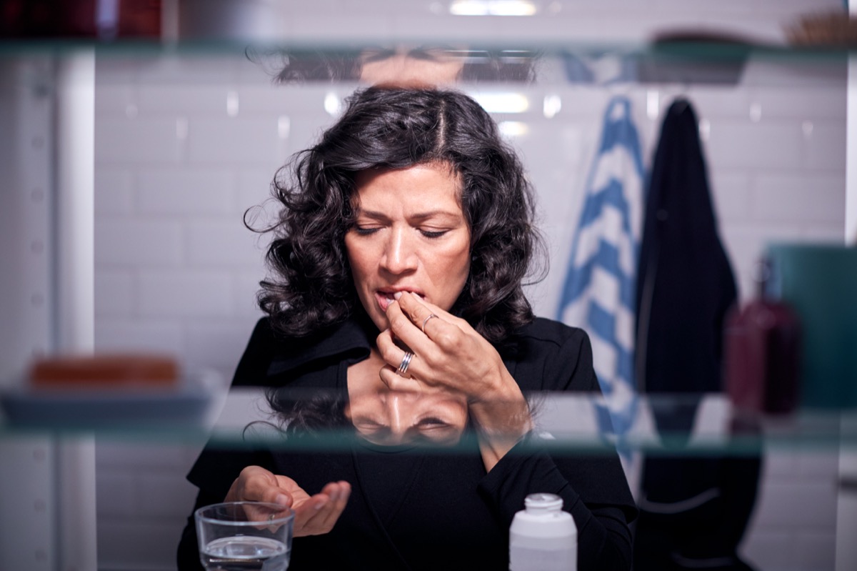 Vedere prin dulapul de baie a unei femei mature care ia medicamente cu un pahar cu apă