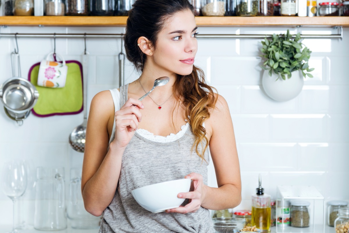 young woman in gray tank top eating from white bowl in modern kitchen