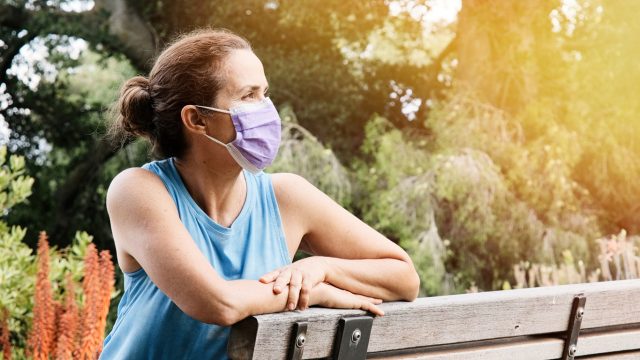 A woman sitting on a park bench while wearing a face mask.