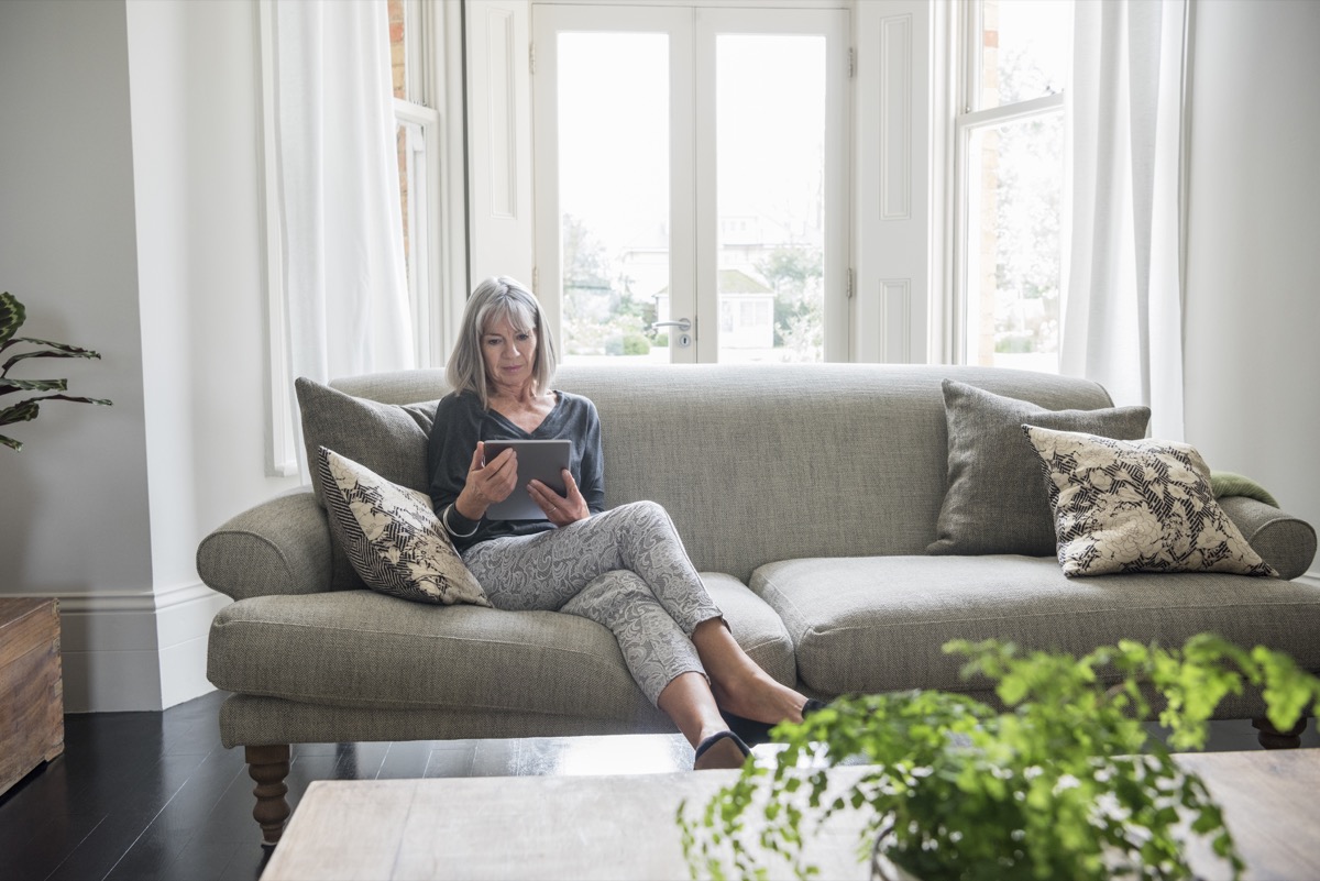 Woman in her 60s at home using a tablet. She is sitting on a grey sofa in front of a window in the living room, an dis comfortable and relaxed.