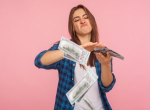 Portrait of wasteful rich girl in checkered shirt scattering dollars with arrogant grimace, boasting wealthy life, concept of careless money spending. indoor studio shot isolated on pink background