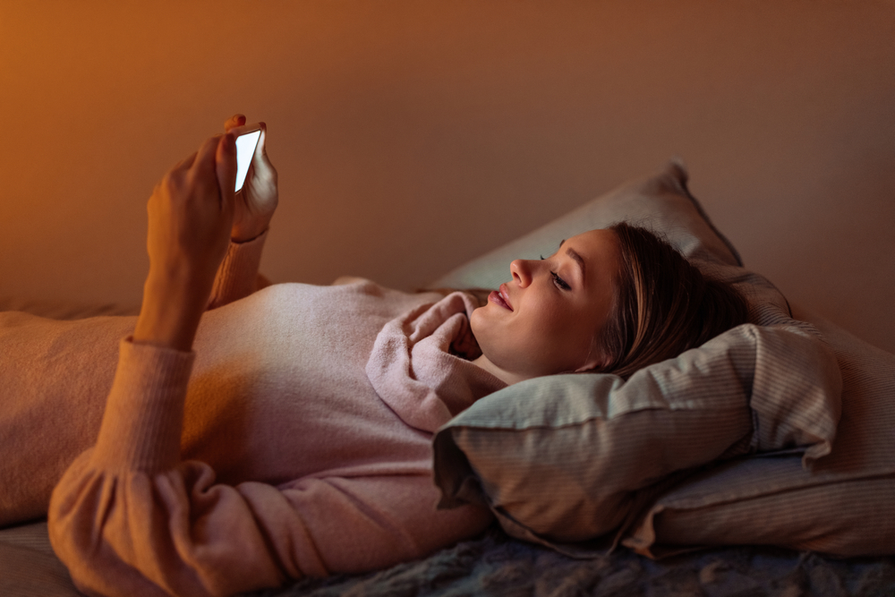 Млада жена, лежаща в леглото, докато използва своя смартфон