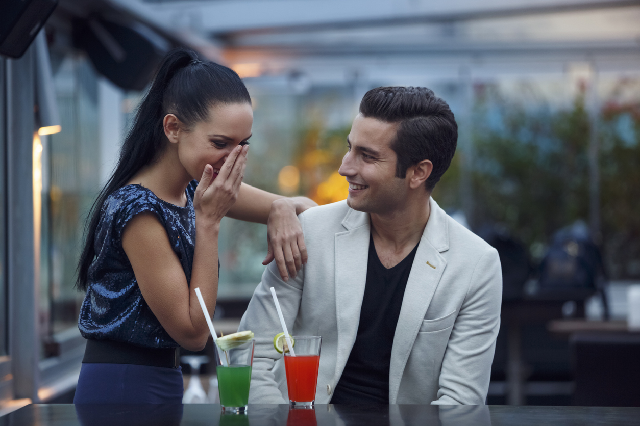 A young man and woman on a date with cocktails in front of them while the young woman covers her mouth