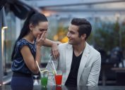 A young man and woman on a date with cocktails in front of them while the young woman covers her mouth