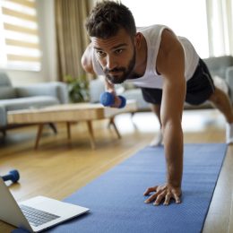 A young man exercising in his living room with his laptop, a yoga mat, and a weight.