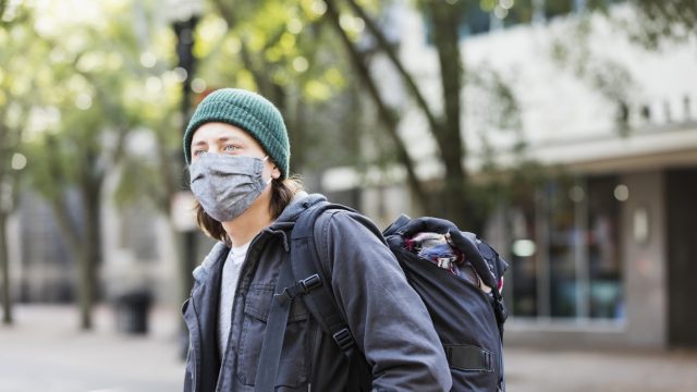 A young man wearing a face mask and a backpack stands on a city street.