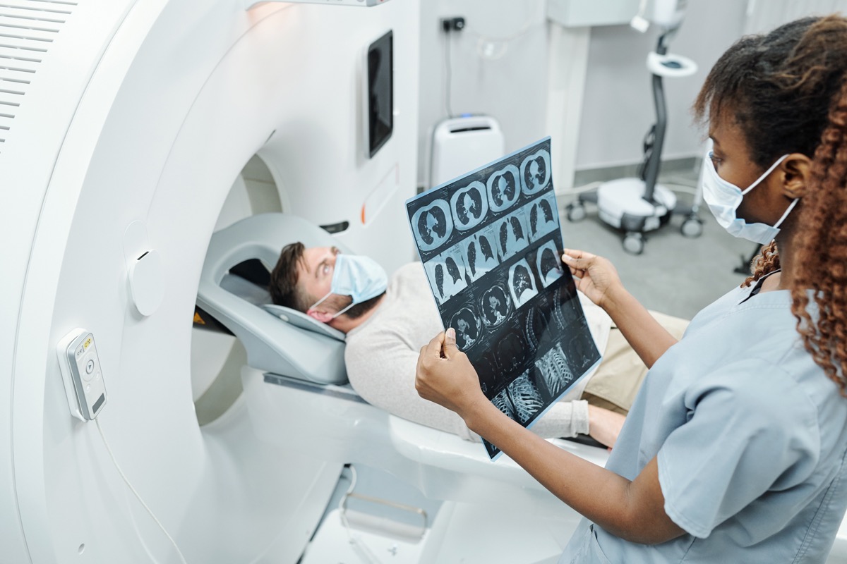 radiologist in uniform and protective mask looking at x-ray image of patient lying on long couchette of medical equipment