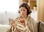 people, health and medicine concept - sad sick young woman in blanket pouring antipyretic or cough syrup from bottle to spoon at home