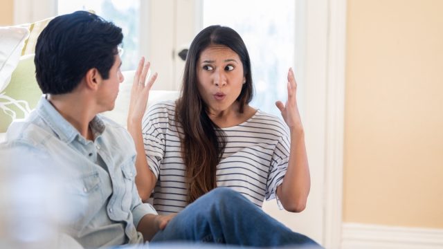 An animated mid adult woman throws up her hands in frustration as she speaks to her unrecognizable husband at home. They are sitting on their living room floor.