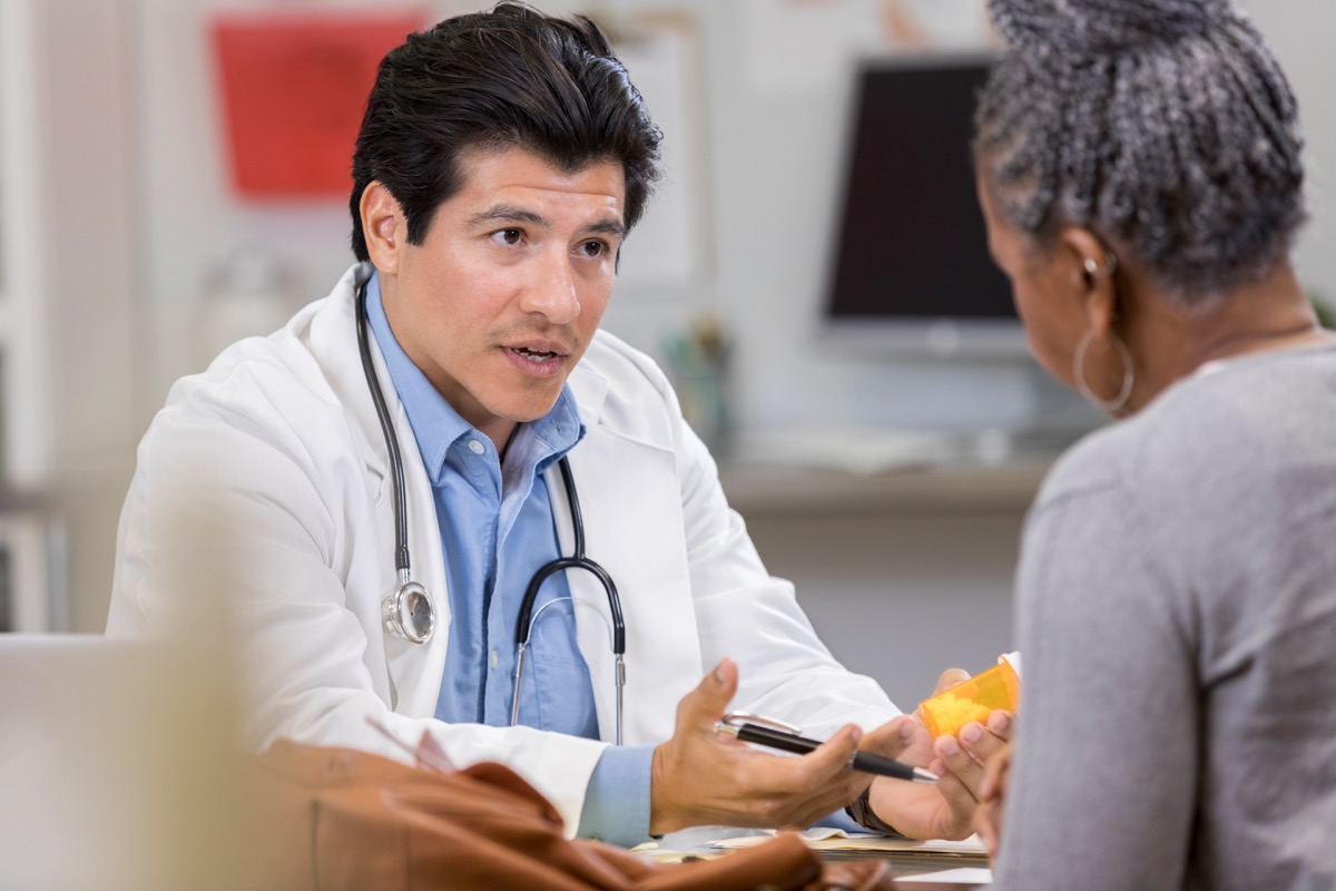 A confident male doctor sits across from an unrecognizable female patient and holds a medication. He gestures as he explains the new prescription.