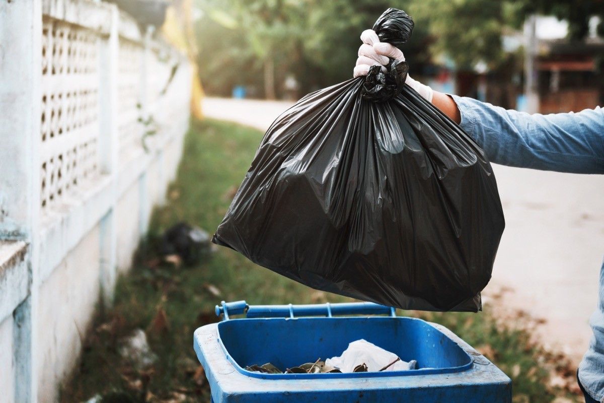 person putting a black garbage bag in a large outdoor garbage can