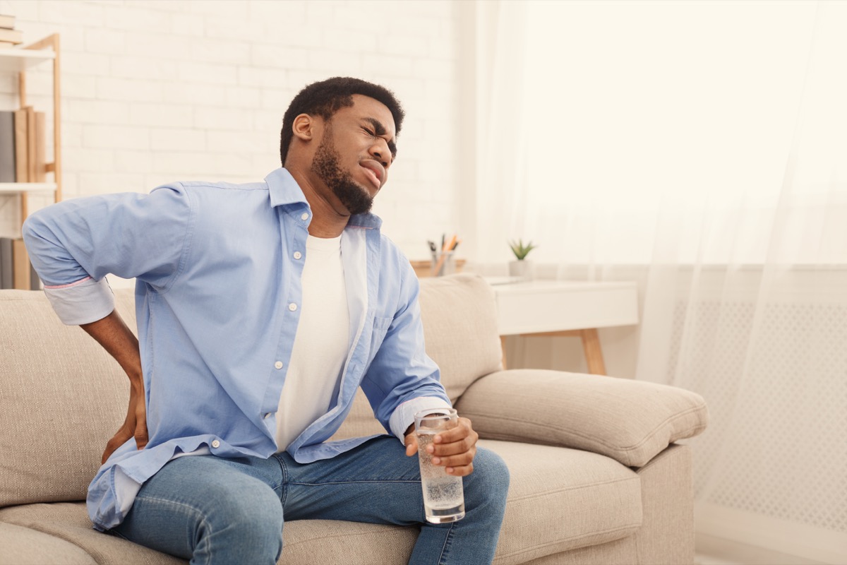 man with back pain, pressing on hip with painful expression, sitting on sofa at home with glass of water, copy space