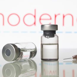 Two vials of vaccine and a syringe in front of the Moderna logo
