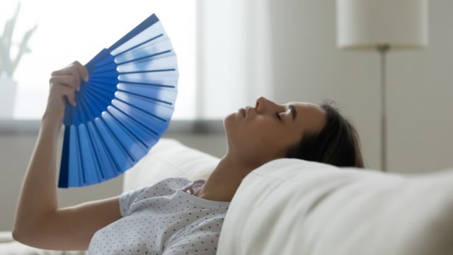 Woman puts head on sofa cushions closed eyes feels sluggish due unbearable heat, waves hand blue fan cool herself