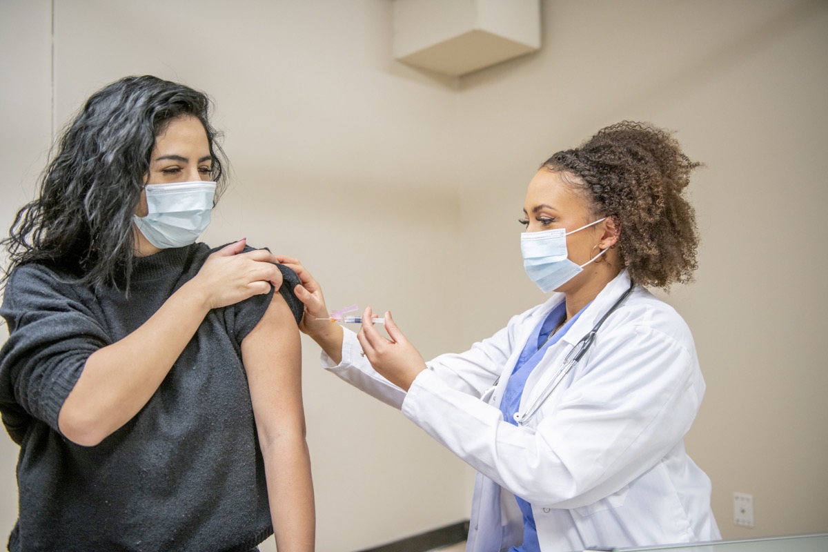 A young female adult is at her doctor's office to receive the COVID-19 vaccination. She is holding the sleeve of her shirt up and her doctor is preparing to administer the shot onto her arm. They are both wearing a face mask to protect themselves from germs.