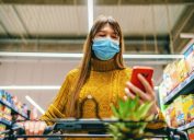 A young woman wearing a face mask looks at her phone while grocery shopping