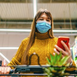 A young woman wearing a face mask looks at her phone while grocery shopping
