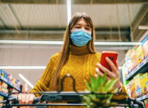 A young woman wearing a face mask looks at her phone while grocery shopping