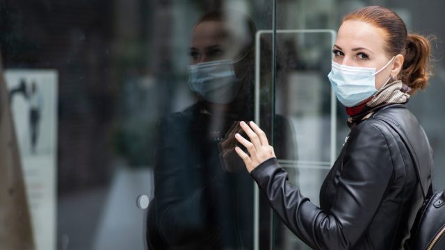 A woman wearing a face mask while looking inside a closed store with a sad look on her face