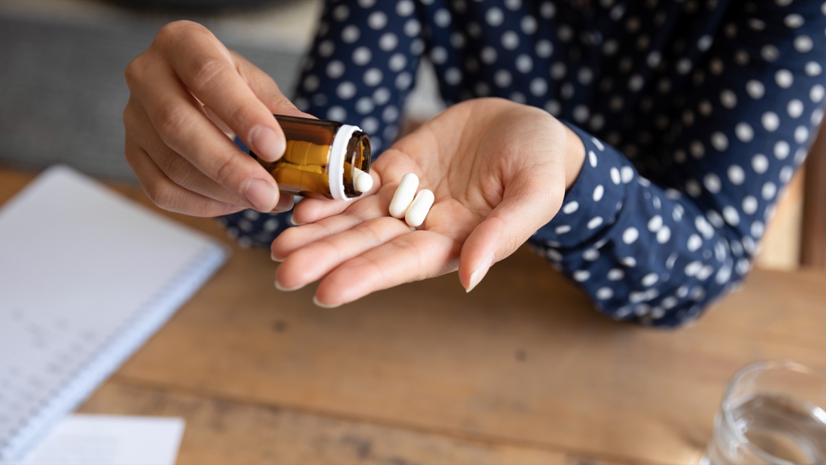 Close up young woman pouring pills out of bottle. Stressed millennial student holding aspirin painkiller antidepressant antibiotic to relieve pain, feeling unhealthy at home or office.