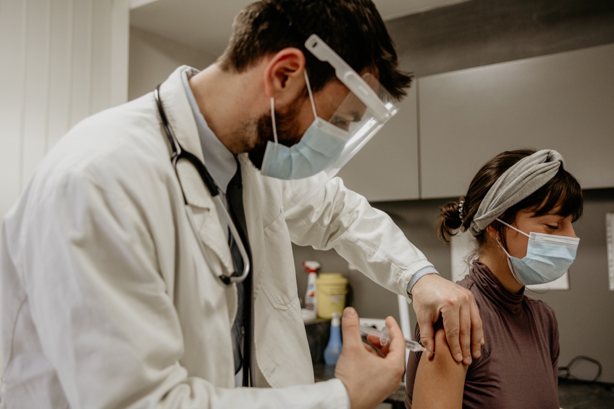 Photo of a male doctor wearing his protective workwear, giving a vaccine to his patient in the clinic . She is wearing a protective face mask as well, looking down