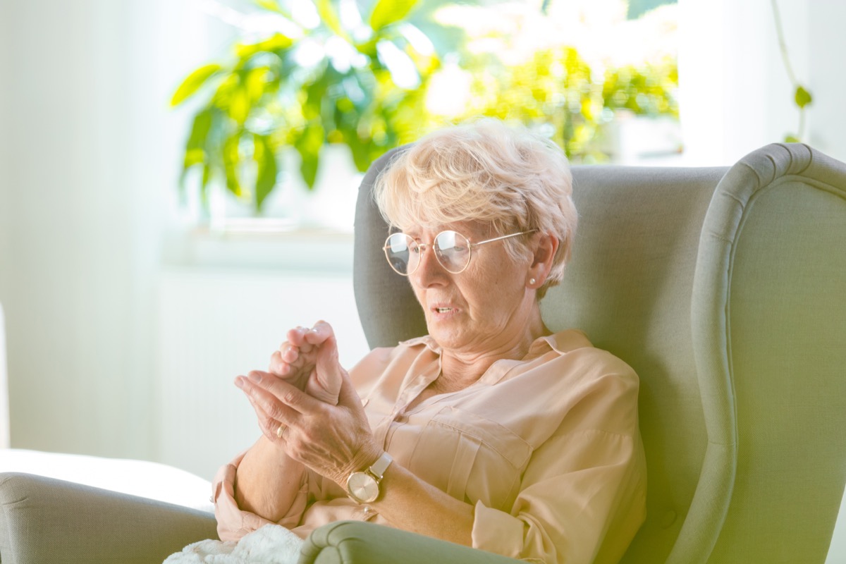 Elderly woman suffering from pain in hand at home, she is sitting in armchair in her room