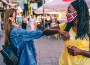 Two young female friends wearing face masks bump elbows to say hello