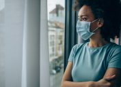 woman wearing protective face mask and looking through window at home during Coronavirus/COVID-19 pandemic.