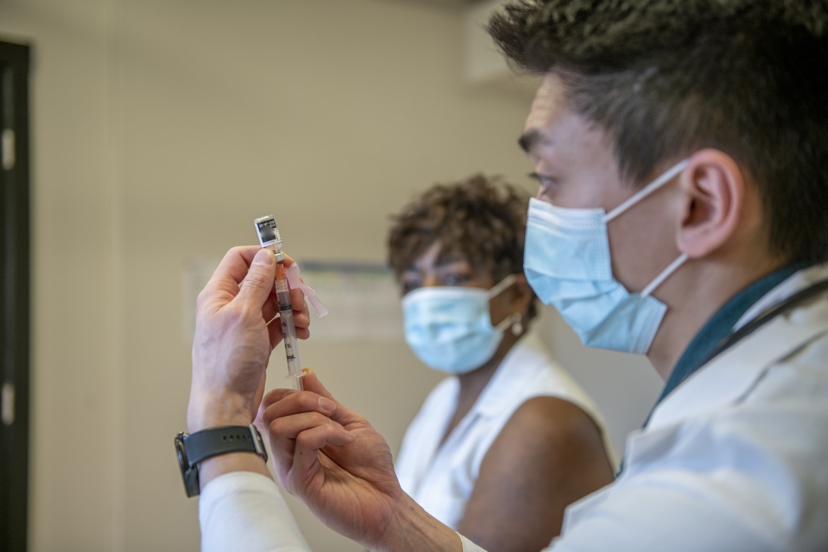 A male healthcare worker is holding a syringe and measuring the right amount of vaccine dosage prior to administering the medical injection onto a patient's arm