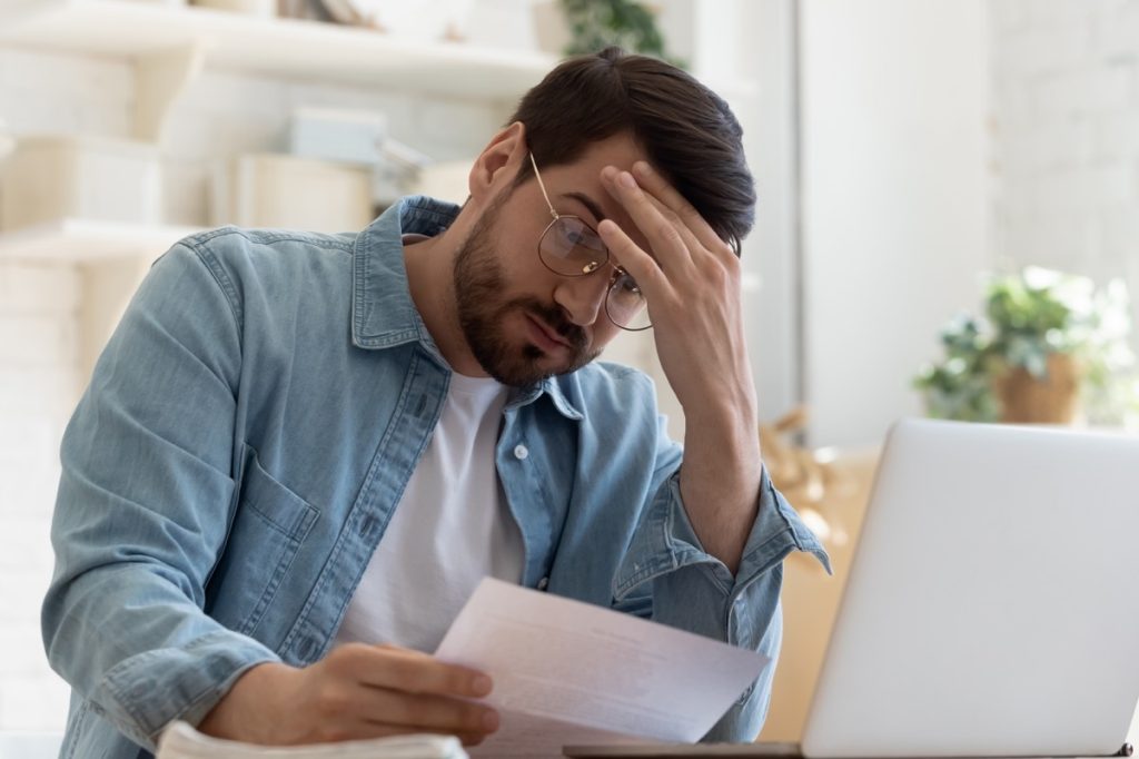 Upset frustrated young man reading bad news in postal mail letter paper document sit at home table, depressed stressed guy worried about high bill tax invoice, overdue debt notification money problem