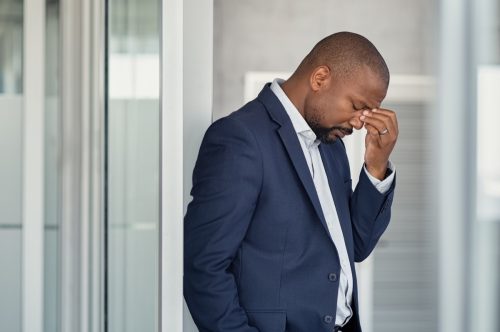 Stressed mature business man rubbing eyes standing in office. African american businessman in formal clothing feeling tired rubbing nose and eyes, for fatigue and headache. Depressed and anxious man in office feeling frustrated after layoff.