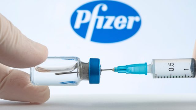 A set of hands filling a syringe from a vial in front of the Pfizer logo